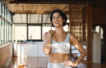 Woman in athletic wear smiles in a sunlit room, standing with one hand on her hip. Background features large windows and wooden walls.