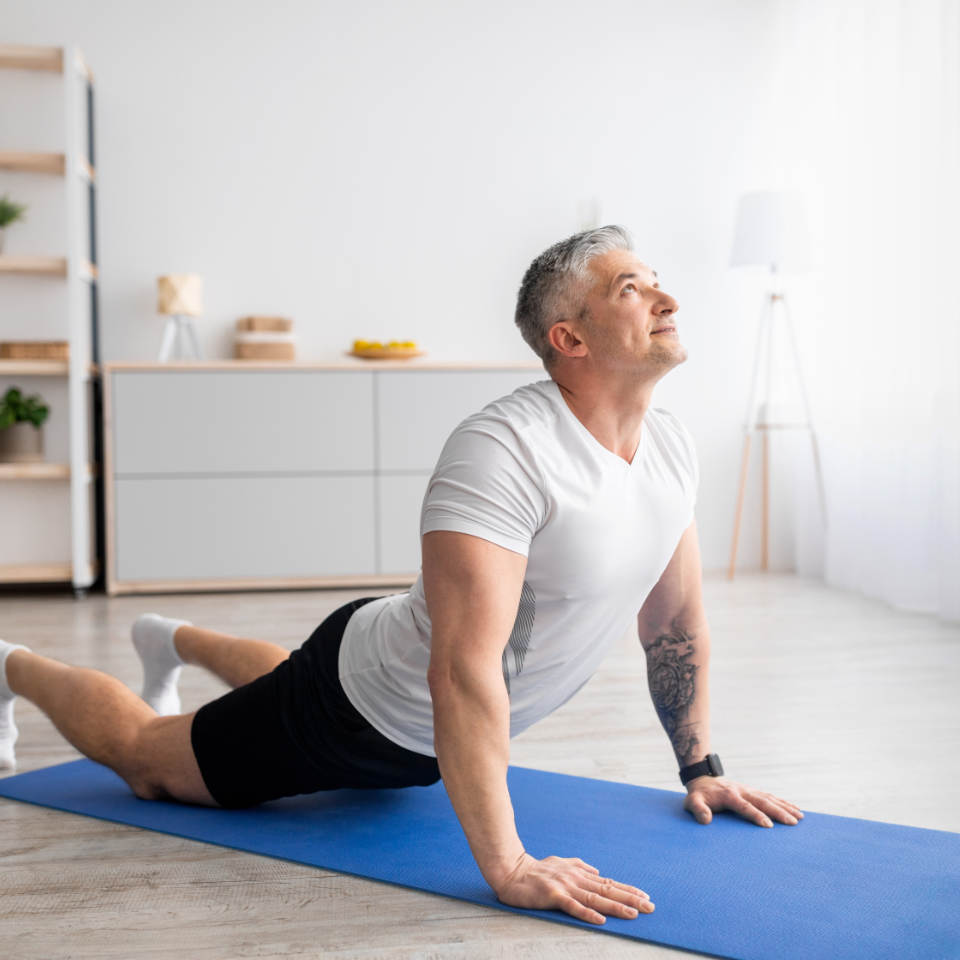 Middle-aged man doing an upward-facing dog yoga pose on a blue mat in a bright, modern living room.