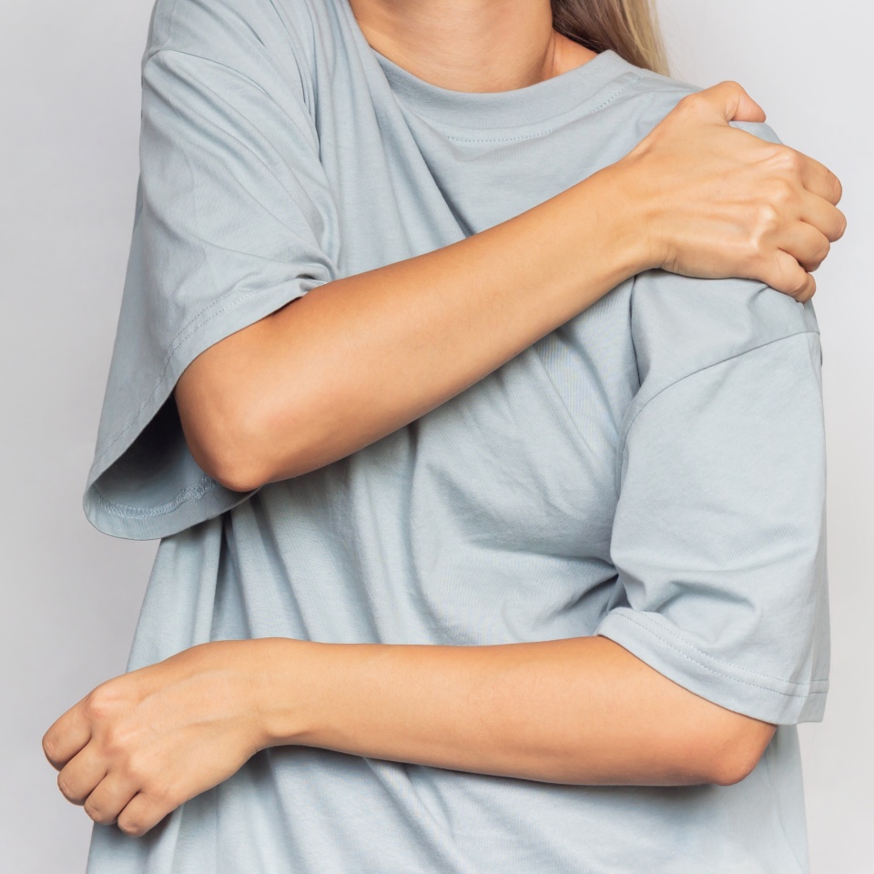 Cropped shot of a young woman in a gray t-shirt holding a shoulder in her hands isolated on a grey background. Shoulder injuries, arm pain, neuralgia