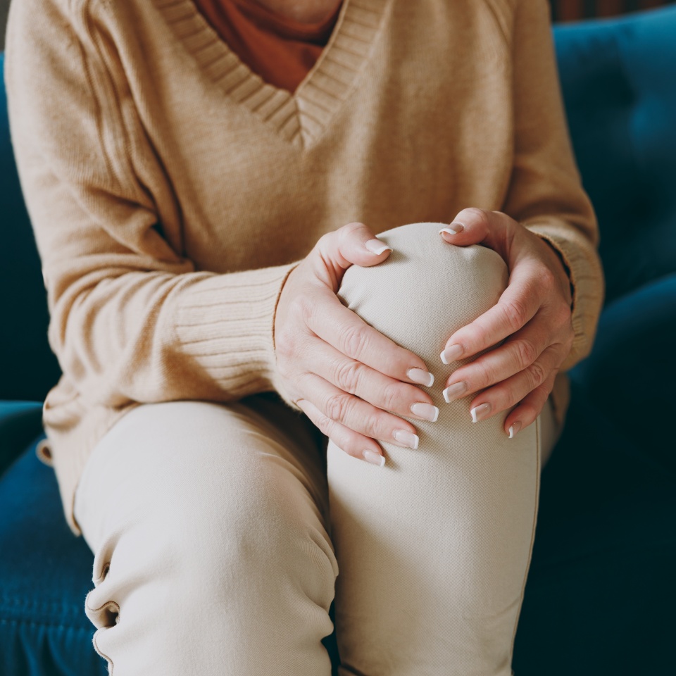 Cropped close up photo of pensioner woman wear casual clothes sits on blue sofa hold knee suffer from leg knee pain inflammation stay home flat spend free spare time in living room indoor grey wall