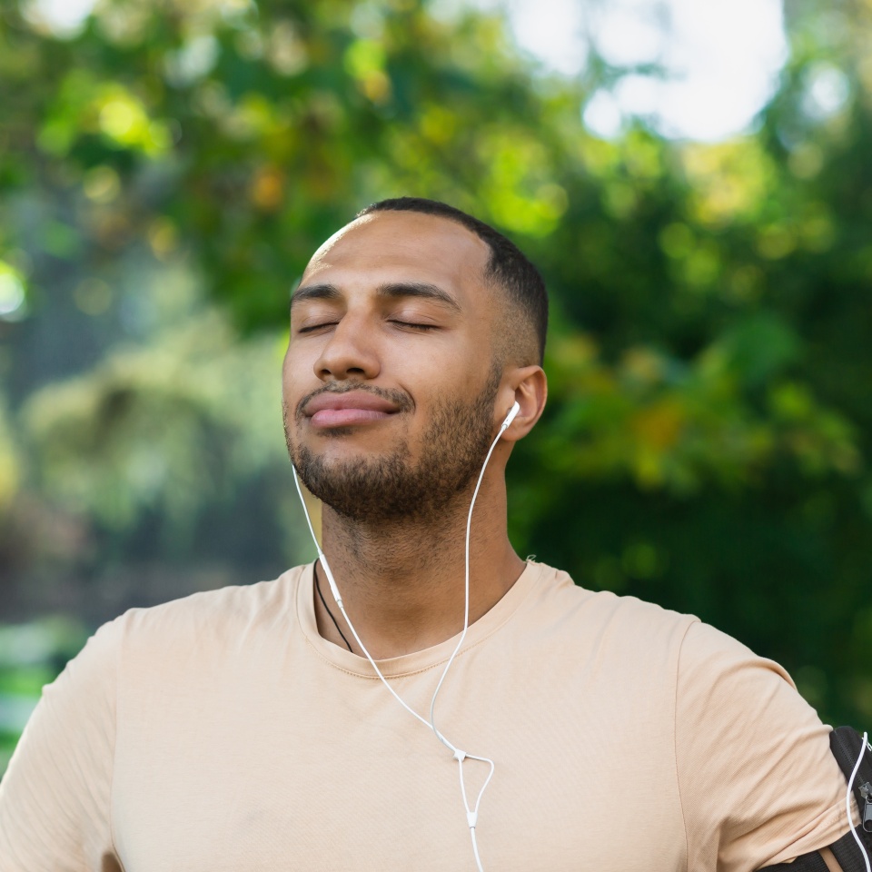 Close-up portrait of sportsman in park, hispanic man jogging in park with eyes closed breathing fresh air and resting, jogging with headphones listening to music and online radio and podcasts.