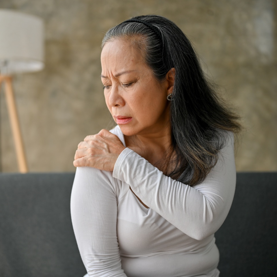 Unhappy Asian mature woman suffering from shoulder pain, massaging her shoulder while resting on sofa in her living room.