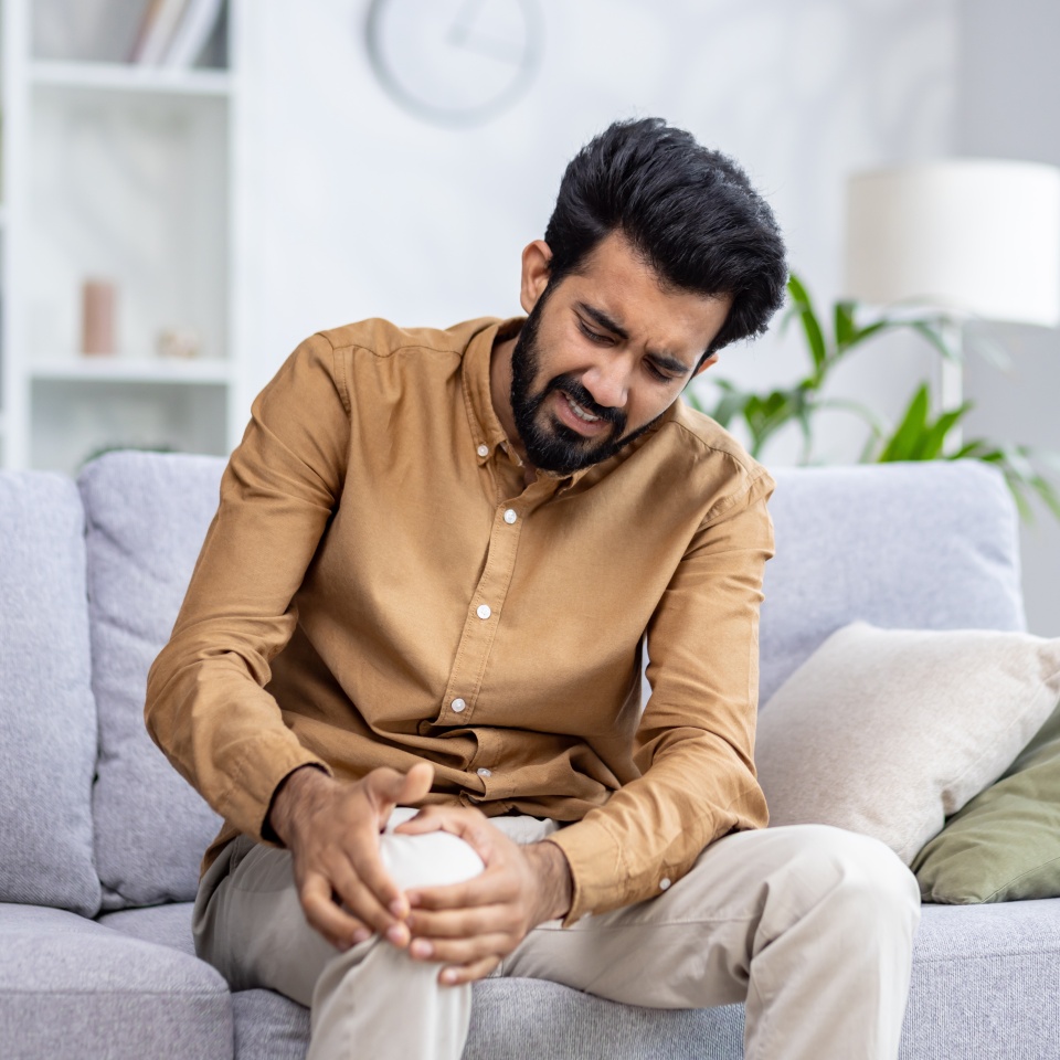 Indian young man sitting on the sofa at home and holding his knee, suffering and grimacing in pain.