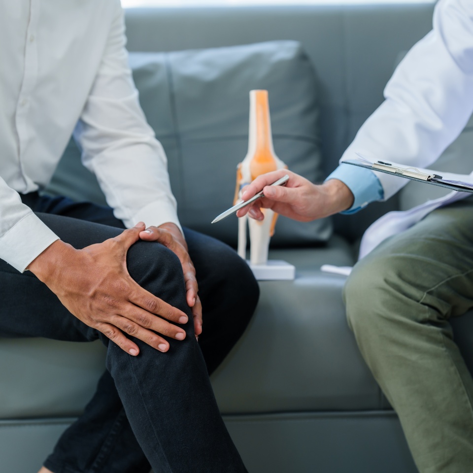 Doctor is seeing a patient. sitting and holding his knees with discomfort Meanwhile, bone model. talking about knee pain or osteoarthritis.