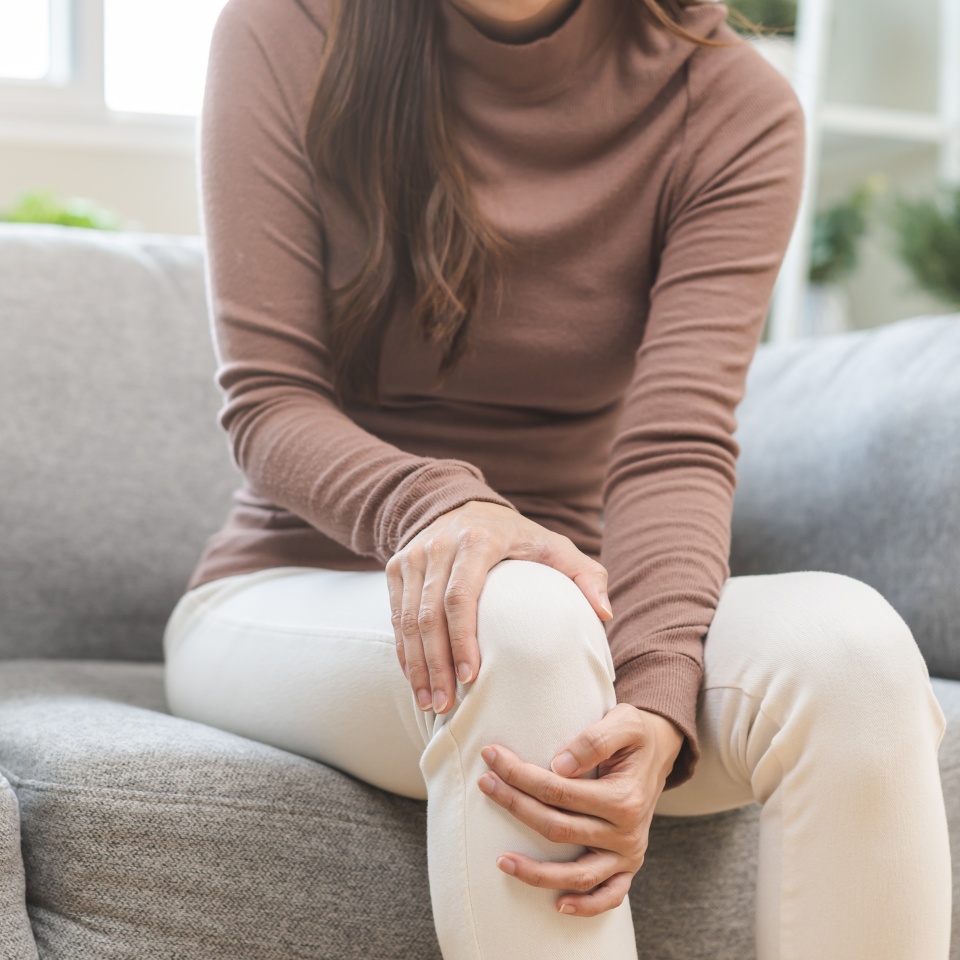 Health care of knee pain concept, close up hand of woman arthritis sitting joint ache, sore cramp or sprain tendon in leg on sofa at home. Osteoarthritis chronic disease of body problem from trauma.