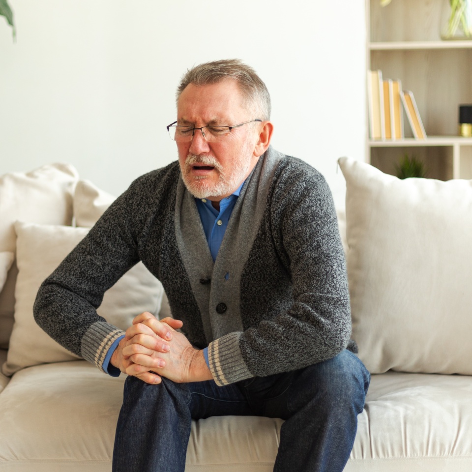 Knee pain arthritis body sick health care concept. Unhappy middle aged senior man suffering from knee ache sitting on sofa at home. Mature old senior grandfather touching leg feeling pain hurt in knee