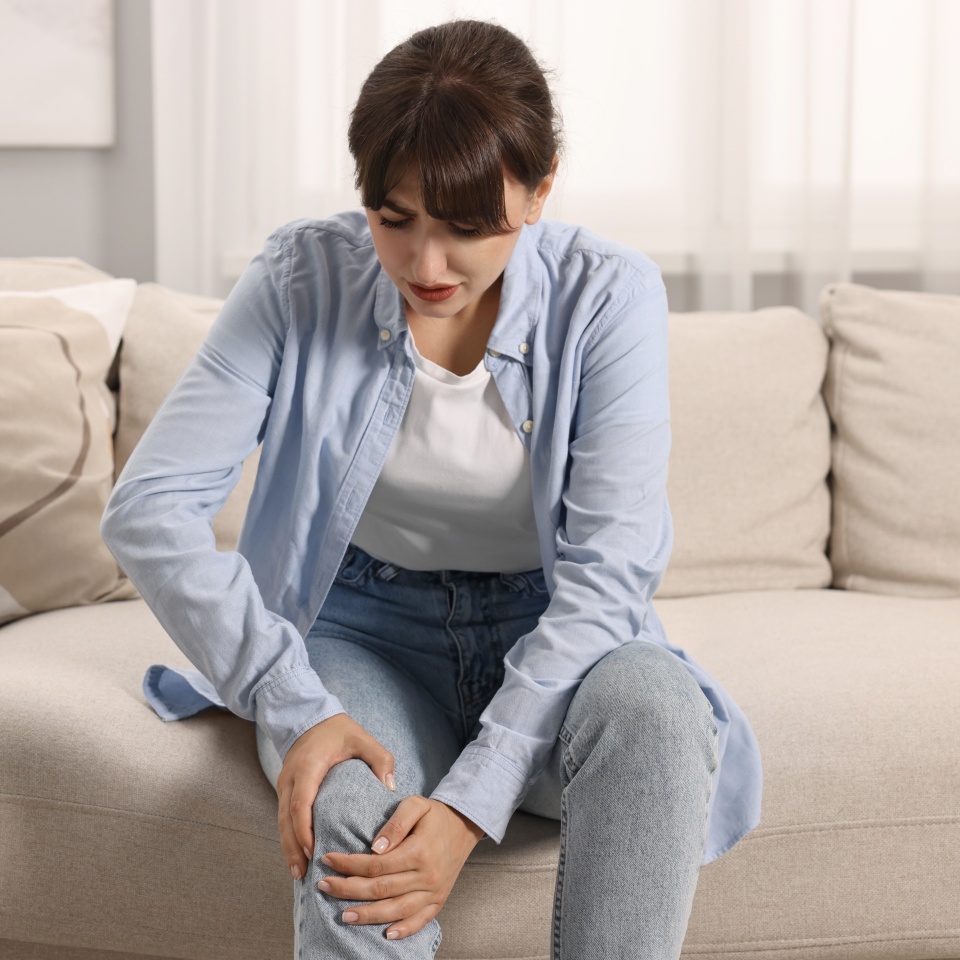 Upset woman suffering from knee pain on sofa at home