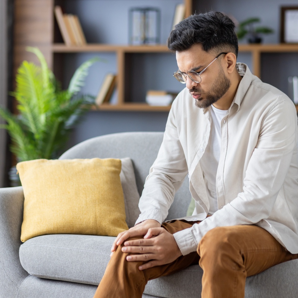 Contorted in pain, a young Indian man sits on the sofa at home and holds his knee with his hand, feels severe pain in his leg.