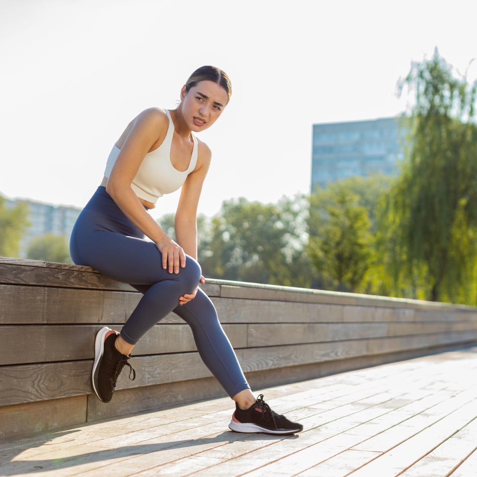 Young sportswoman sitting with knee pain in city park. Woman massaging stretching, trauma injury while jogging outdoors. Fitness female sprain severe pain stretch pull. Leg muscle cramp calf sport.