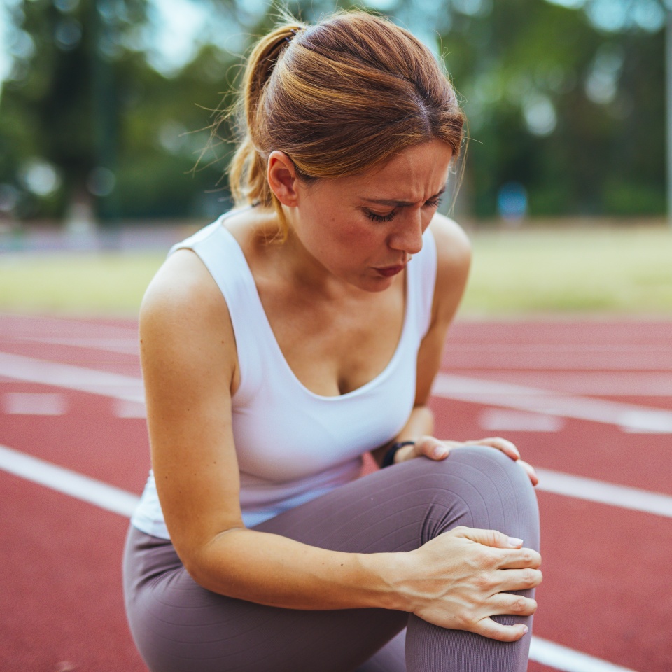A woman experiencing leg pain while kneeling on an athletic track. She is holding her knee, indicating possible injury or strain. The outdoor setting suggests a focus on sports and fitness activities.