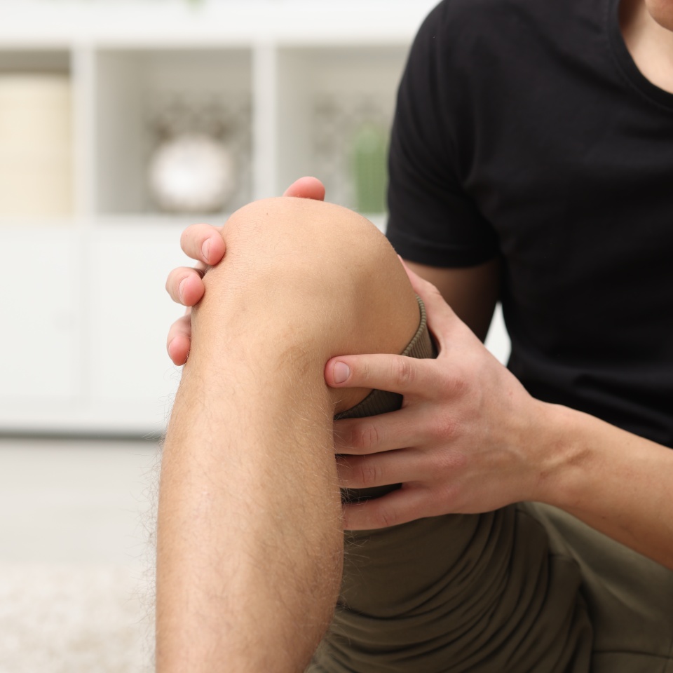 Young man suffering from pain in knee at home, closeup. Space for text