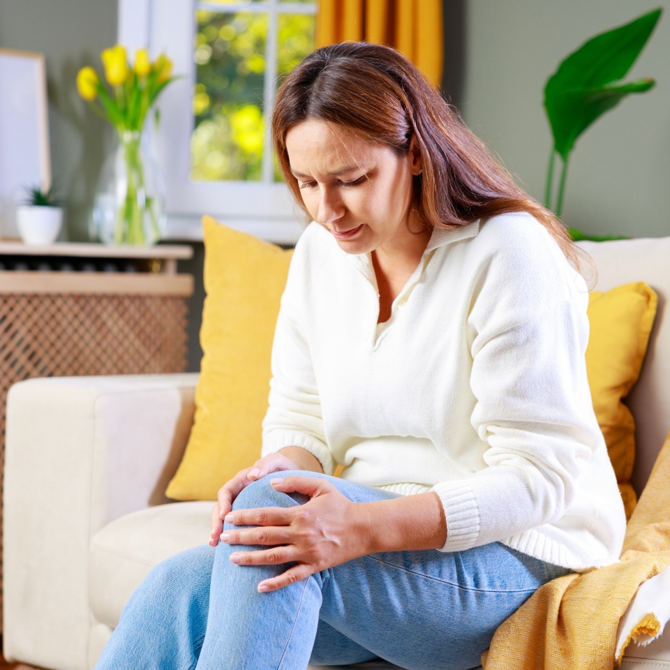 Young woman suffering from pain in knee. Mid adult age woman feeling unwell, suffering from pain in kneee while sitting on sofa in the living room at home