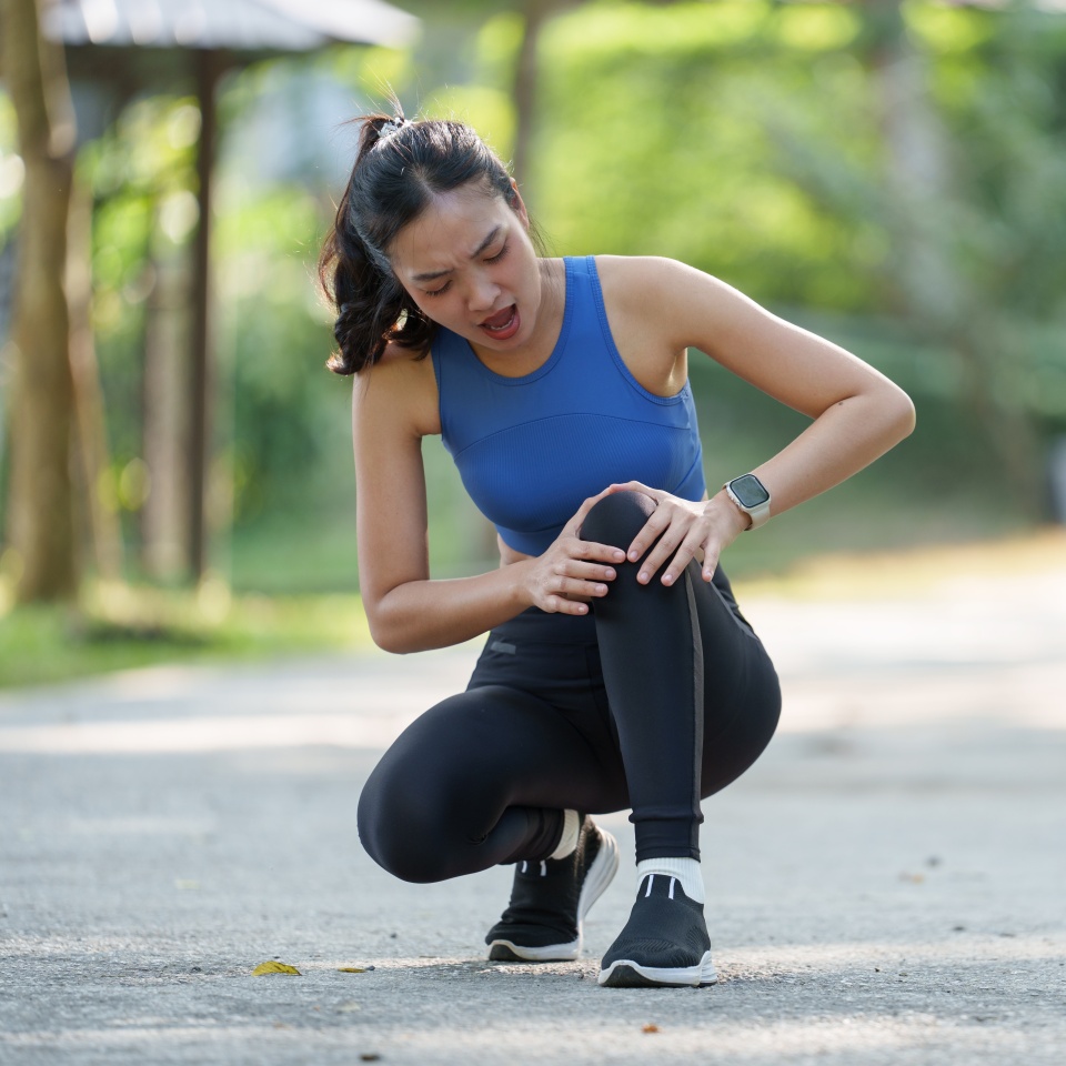 Young sporty asian woman experiencing a painful knee injury during an outdoor workout, gripping her leg in discomfort and searching for relief amid her fitness routine