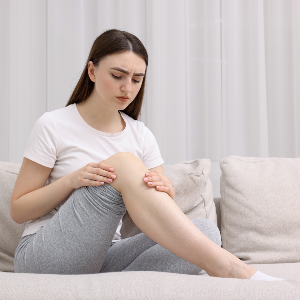 Young woman suffering from pain in knee on sofa at home. Space for text