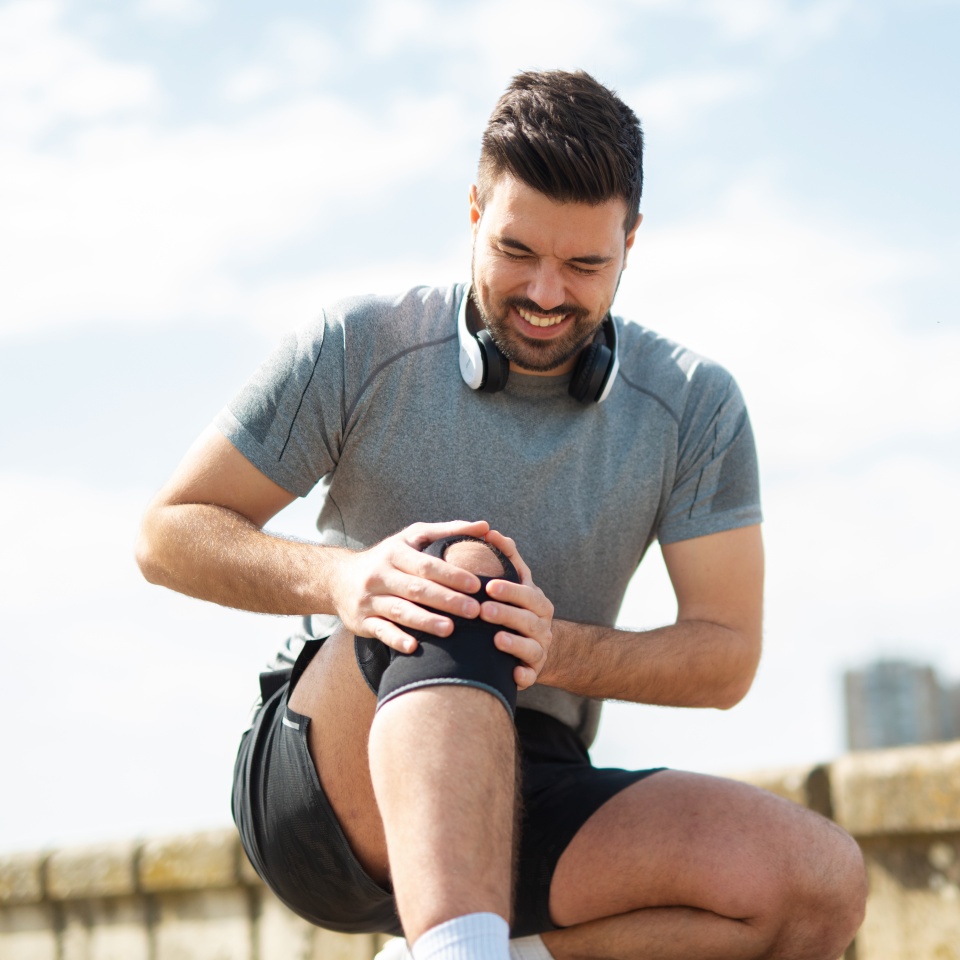 Young man outdoors holding his knee in pain after exercise, wearing sportswear and headphones, suffering from injury or joint discomfort.