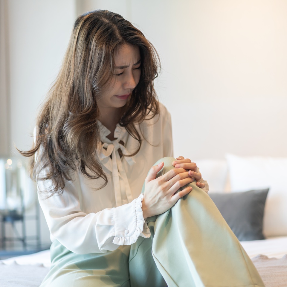 Asian woman suffering from knee pain and inflammation, arthritis symptoms sitting on sofa, health and wellness, joint pain, lifestyle health concept at home, healthcare awareness, knee problem relief