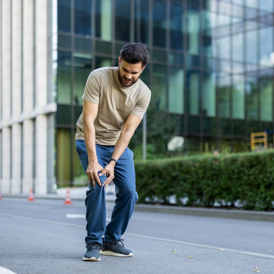 An Indian young man is busy running and exercising outside, injured, standing crooked in pain and holding his knee.