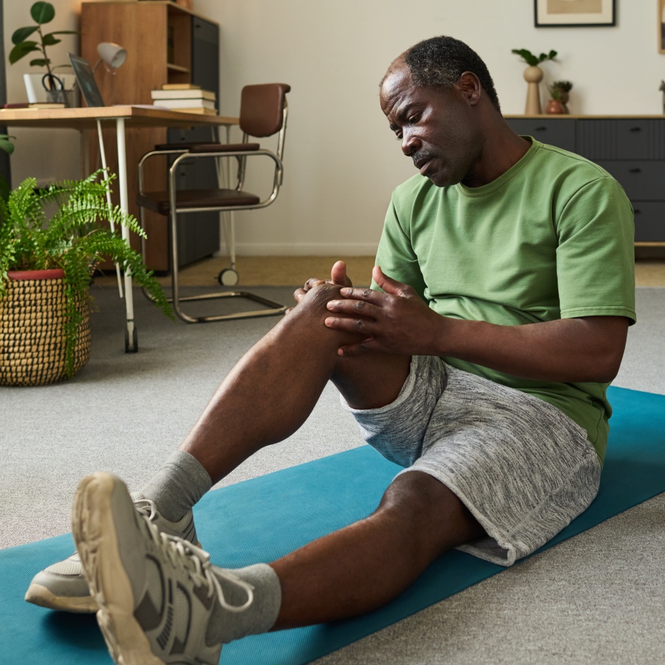 Senior Black man sitting on exercise mat holding knee with both hands showing discomfort during home workout session indoor, fitness equipment and plants visible in background