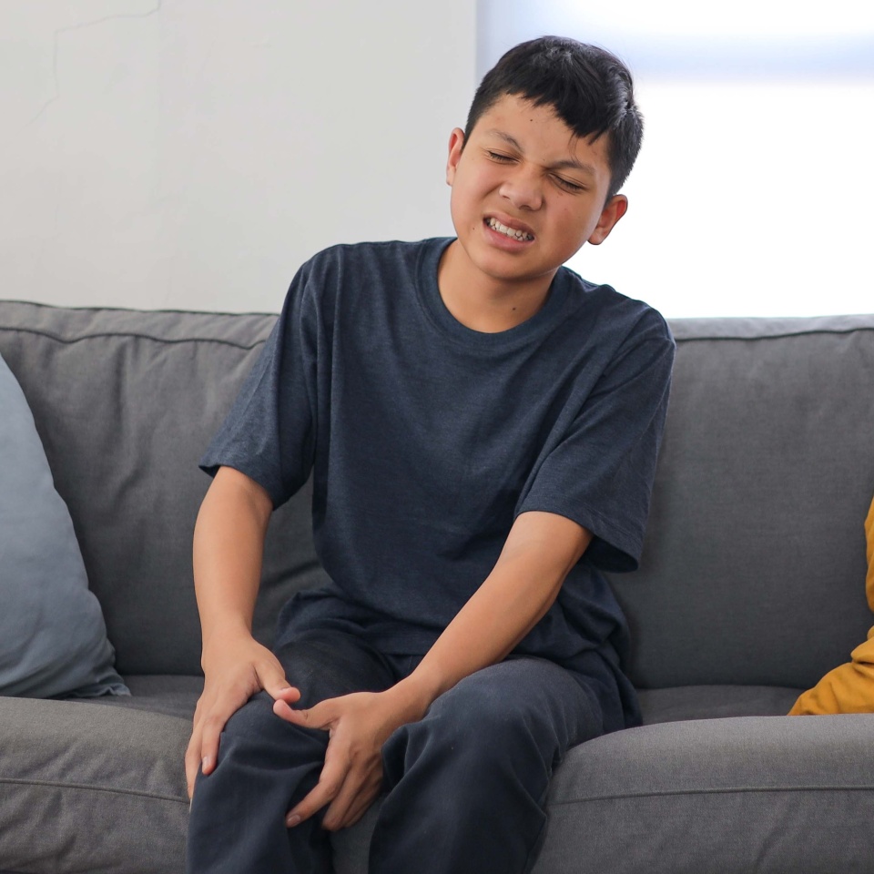 Asian Kid Boy Suffering from Knee Pain Injury While Seating on Sofa in Living Room