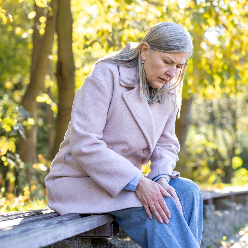 Elder woman sitting on a park bench experiencing acute knee pain, holding her leg in discomfort due to health issues, joint pain, or arthritis in an outdoor setting