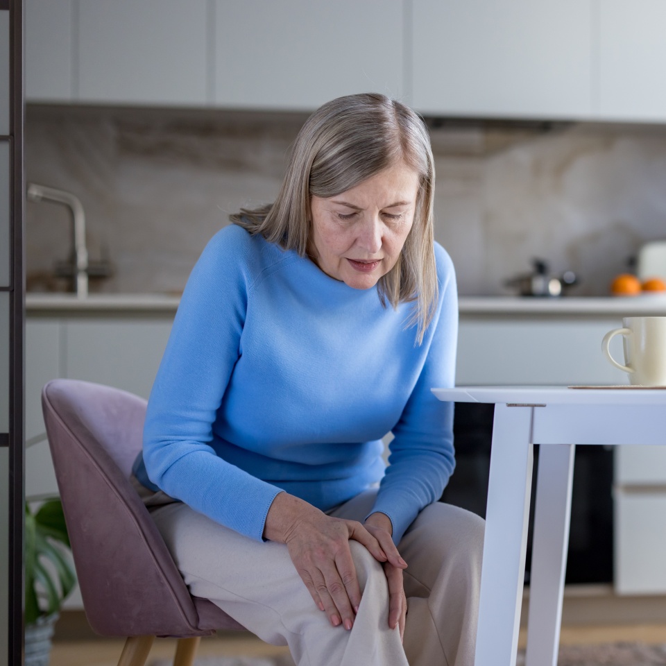 Senior woman sitting in her kitchen gripping her painful knee, showing discomfort from age-related joint pain and arthritis affecting everyday home life and mobility
