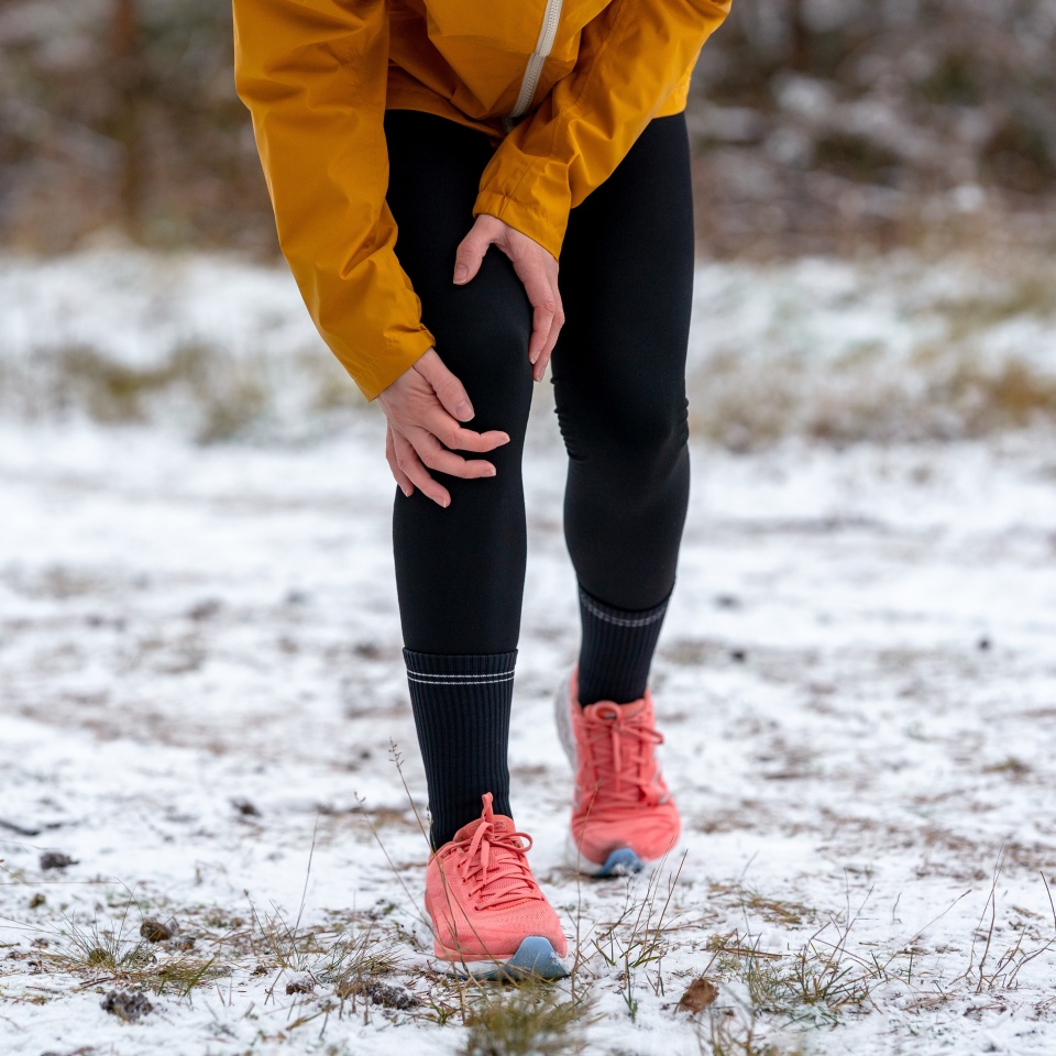 Close-up of a female runner clutching her knee, having stopped on a snowy trail. Sports injury or sudden training setback in cold winter weather.